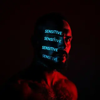 @text1: Retrato en clave baja de un hombre rudo con bigote y piercing facial, iluminado con luz dura y direccional que crea sombras intensas y alto contraste. Fondo profundo en tonos rojo oscuro y negro. La palabra "SENSITIVE" se proyecta repetidamente en una luz fría azul, cruzando parcialmente el rostro y torso, mientras grandes áreas permanecen en penumbra, generando una atmósfera cinematográfica íntima, masculina pero vulnerable. Estilo de fotografía artística contemporánea, con amplias zona, photorealistic style, detailed texture, high resolution photography, professional lighting